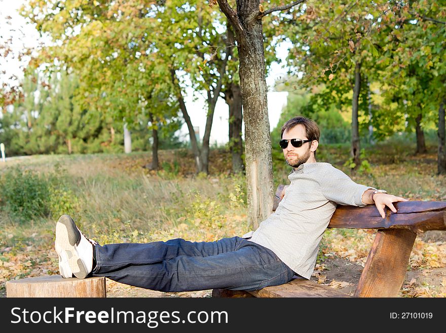 Young man relaxing in the sun sitting on a rustic wooden bench with his feet up on a tree stump with copyspace