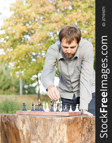 Man standing over a chessboard on a rustic wooden tree stump table playing chess outdoors. Man standing over a chessboard on a rustic wooden tree stump table playing chess outdoors