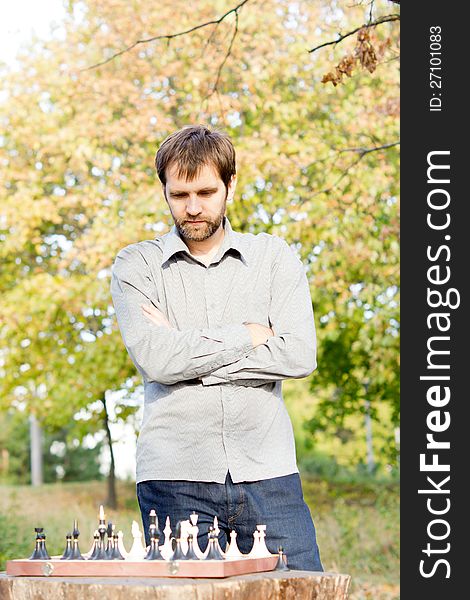 Young bearded man standing with his arms folded above an outdoor chessboard contemplating his next chess move. Young bearded man standing with his arms folded above an outdoor chessboard contemplating his next chess move