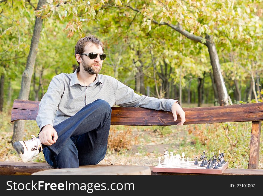 Pensive young man in sunglasses sitting on a bench in woodland with a chessboard alongside him staring off into the distance deep in thought. Pensive young man in sunglasses sitting on a bench in woodland with a chessboard alongside him staring off into the distance deep in thought