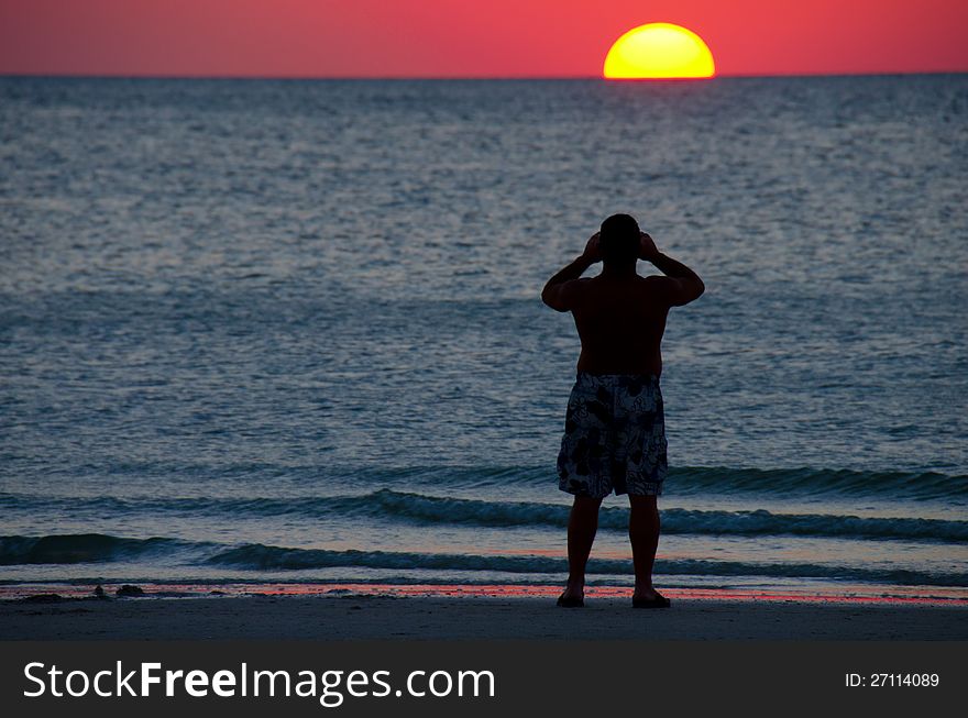 A man standing at the water's edge photographing a colorful sunset at a calm ocean. A man standing at the water's edge photographing a colorful sunset at a calm ocean