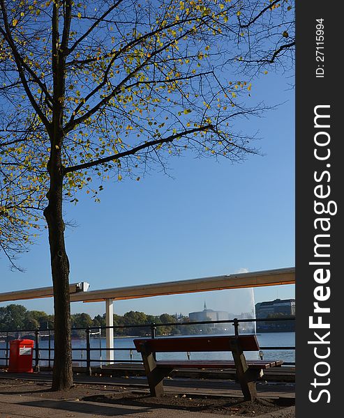 A sunny autumn day in October at the lakefront of the Binnenalster in Hamburg: Leaves are falling over the empty bench and shadows become longer. A sunny autumn day in October at the lakefront of the Binnenalster in Hamburg: Leaves are falling over the empty bench and shadows become longer.