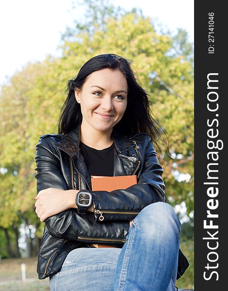 Smiling young casual womanin jeans and a leather jacket holding a book to her chest while sitting outdoors. Smiling young casual womanin jeans and a leather jacket holding a book to her chest while sitting outdoors