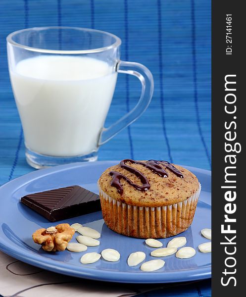 Closeup image of a muffin cake on a blue plate with almond chips, walnut, a piece if dark chocolate and a glass of milk. Closeup image of a muffin cake on a blue plate with almond chips, walnut, a piece if dark chocolate and a glass of milk