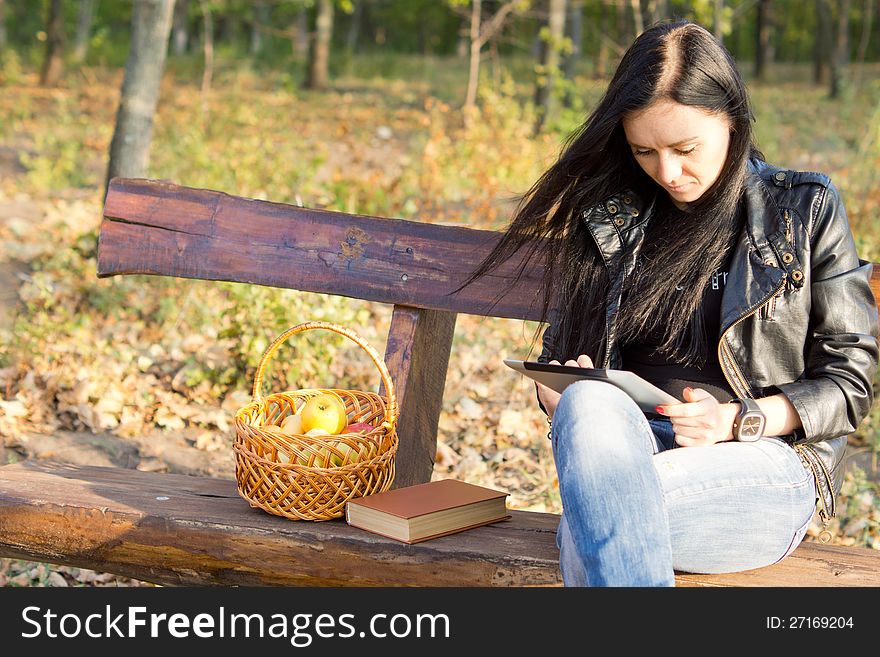 Young casual woman sitting on a rustic wooden park bench with a basket of apples and book using a tablet. Young casual woman sitting on a rustic wooden park bench with a basket of apples and book using a tablet