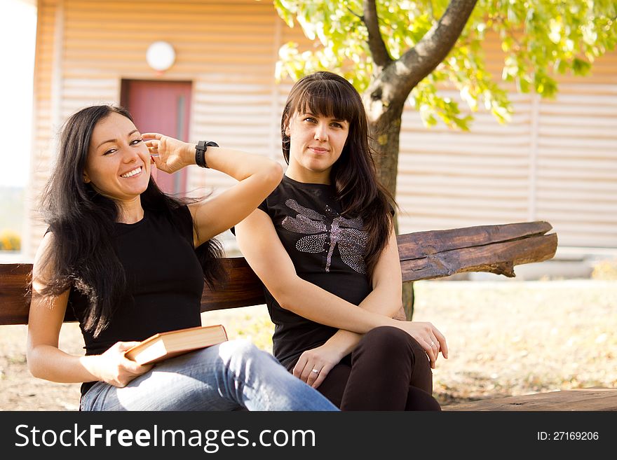 Two attractive young women relaxing and laughing together outdoors sitting on a rustic wooden bench. Two attractive young women relaxing and laughing together outdoors sitting on a rustic wooden bench