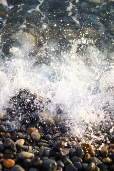 Splashing Waves On The Seashore. The Beach Is Made Of Small Colorful Pebbles. Sea Water Splashes Hitting The Stones. Stock Photo