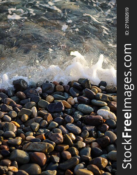 Sea Foam From Small Waves. Crystal Clear Water On The Seashore. The Beach Is Lined With Small Colorful Pebbles.