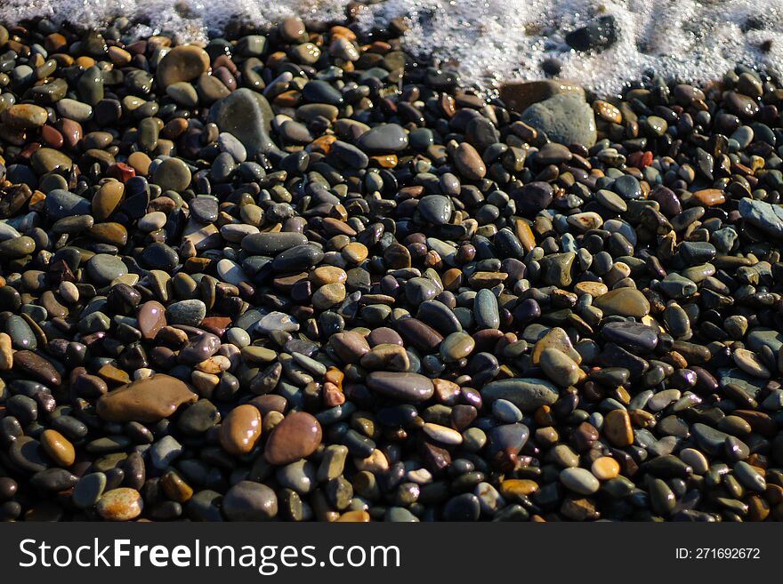 Wet rocks by the sea. The beach is lined with small colorful pebbles. Sea foam from small waves.