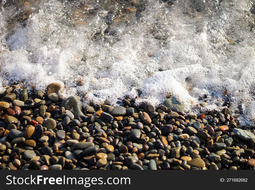 Sea Foam From Small Waves. Crystal Clear Water On The Seashore. The Beach Is Lined With Small Colorful Pebbles.