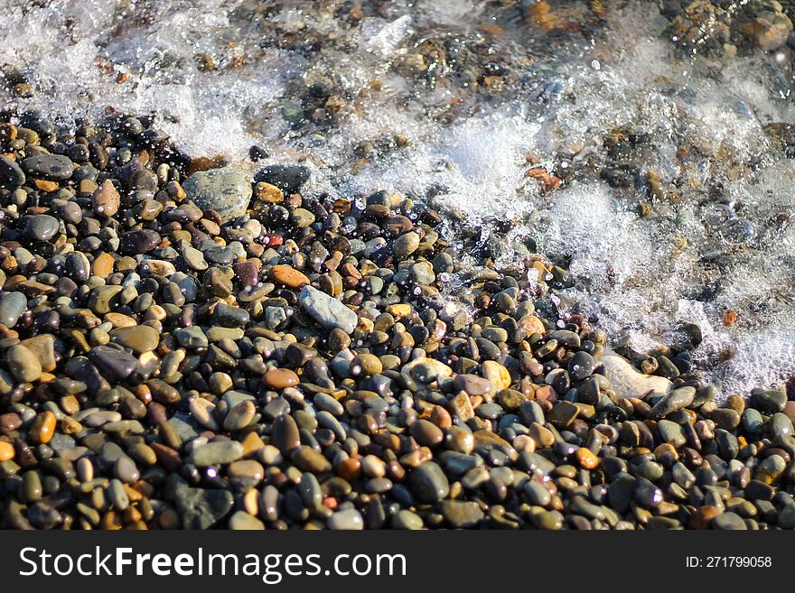 Splashing Waves On The Seashore. The Beach Is Made Of Small Colorful Pebbles. Sea Water Splashes Hitting The Stones.