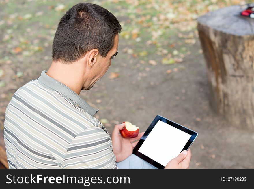 Over the shoulder view of a man sitting on a rural bench using a touchscreen tablet with a blank screen and eating a fresh apple. Over the shoulder view of a man sitting on a rural bench using a touchscreen tablet with a blank screen and eating a fresh apple