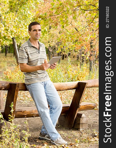 Middle-aged man relaxing on a rustic wooden fence in woodland with a tablet in his hands. Middle-aged man relaxing on a rustic wooden fence in woodland with a tablet in his hands
