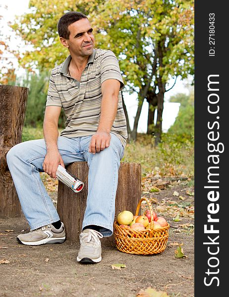 Man seated on a tree trunk in the countryside waitng patiently and looking expectantly off frame with a basket of fresh apples at his feet