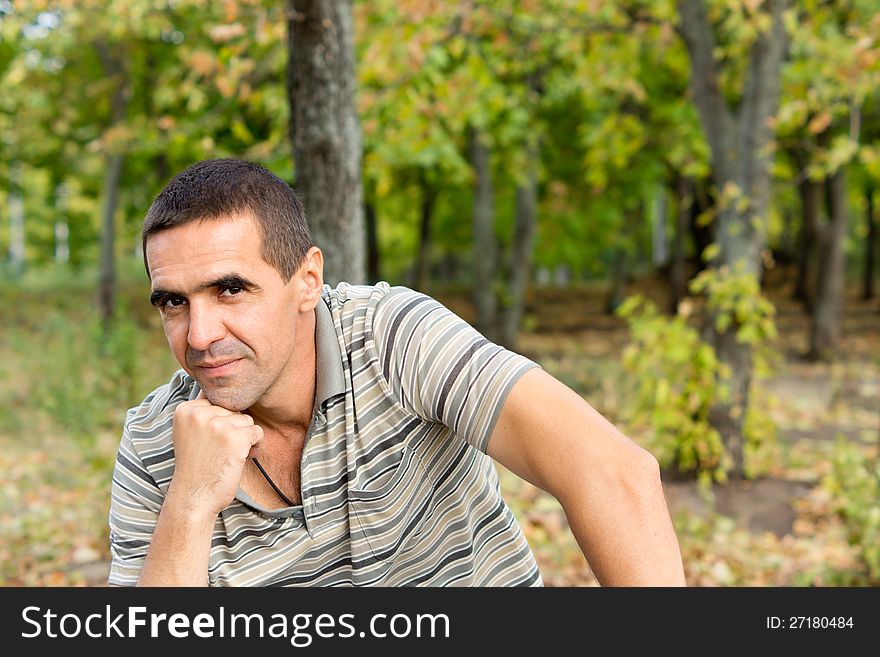 Attracive middle-aged thoughtful man sitting with his chin on his fist giving the camera a slight smile with a woodland backdrop and copyspace. Attracive middle-aged thoughtful man sitting with his chin on his fist giving the camera a slight smile with a woodland backdrop and copyspace