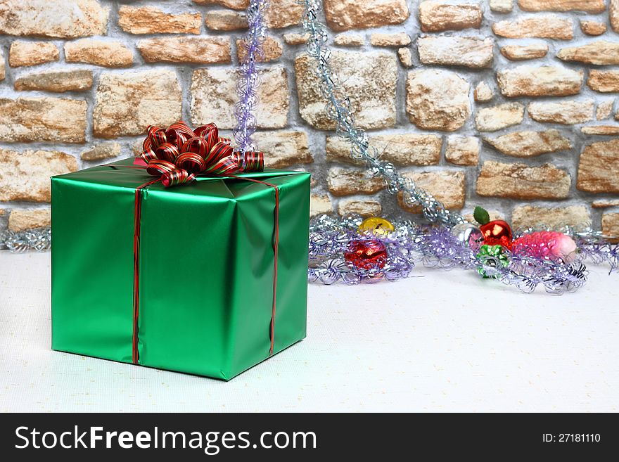 Green satin gift box with ribbon and bow against stone wall with Christmas toys and tinsel in background. Green satin gift box with ribbon and bow against stone wall with Christmas toys and tinsel in background