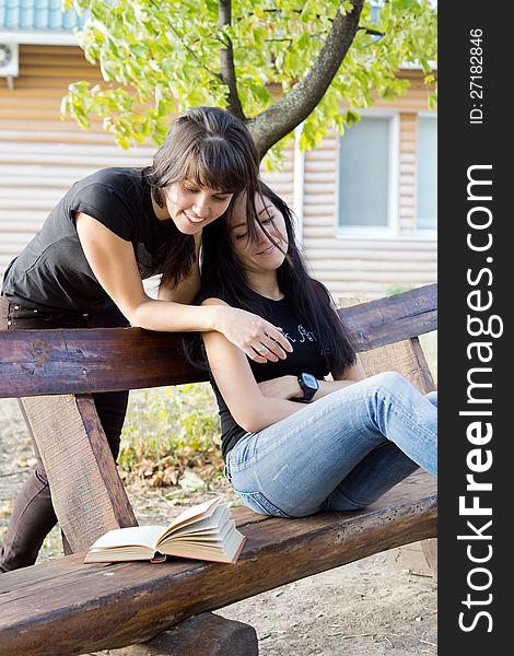 Two young female friends relaxing on a wooden garden bench laughing over a book. Two young female friends relaxing on a wooden garden bench laughing over a book