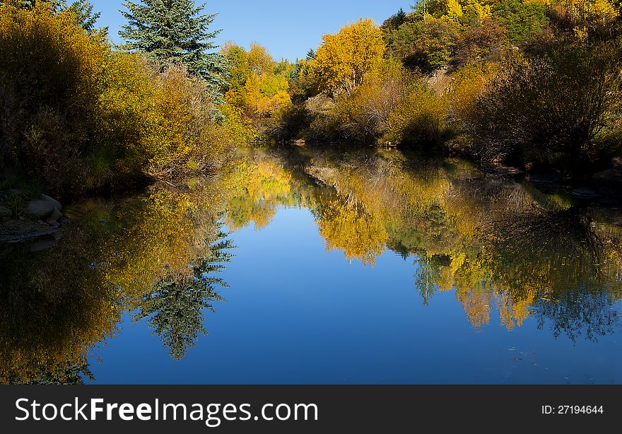 Calm mountain stream surrounded by and reflecting brilliant gold, yellow, orange, green bushes and trees with some brilliant blue sky above and reflecting deep blue in the water. Calm mountain stream surrounded by and reflecting brilliant gold, yellow, orange, green bushes and trees with some brilliant blue sky above and reflecting deep blue in the water.