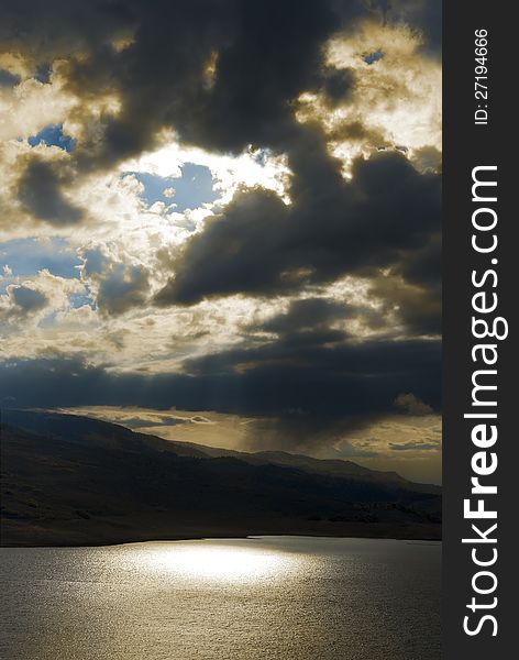 Dramatic stormy sky with sun and blue sky breaking through and light filtering through to hills and a lake glistening below. Dramatic stormy sky with sun and blue sky breaking through and light filtering through to hills and a lake glistening below.