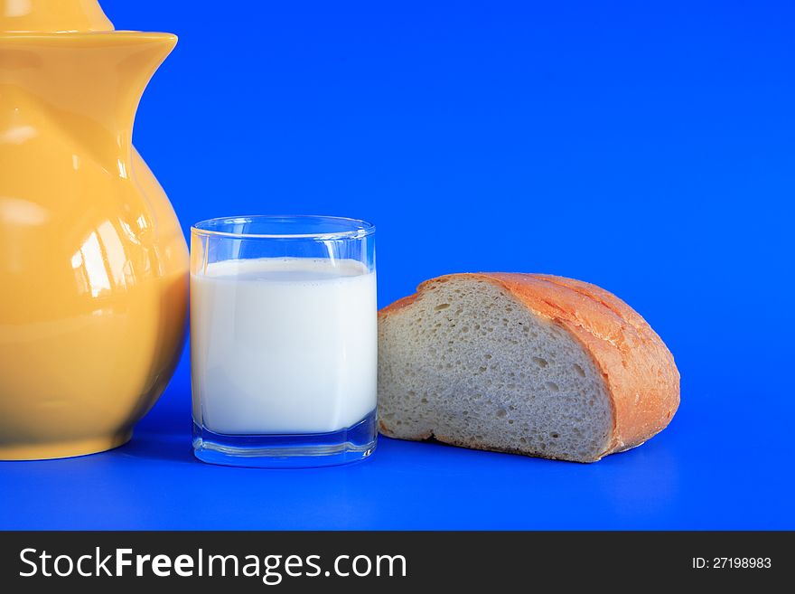 Glass full of milk and bread near yellow pitcher on blue background