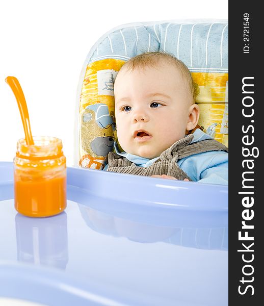 Pretty infant sitting on baby's chair. Looking at orange pulp in jar. White background. Pretty infant sitting on baby's chair. Looking at orange pulp in jar. White background