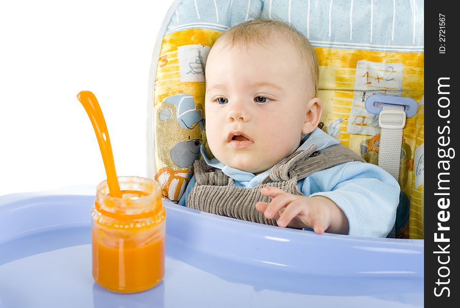 Pretty infant sitting on baby's chair. Looking at orange pulp in jar. White background. Pretty infant sitting on baby's chair. Looking at orange pulp in jar. White background