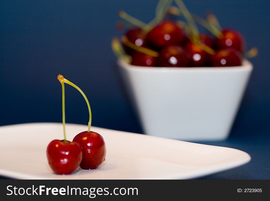 Multiple moist red cherries in a white bowl and 2 cherries in front. Multiple moist red cherries in a white bowl and 2 cherries in front