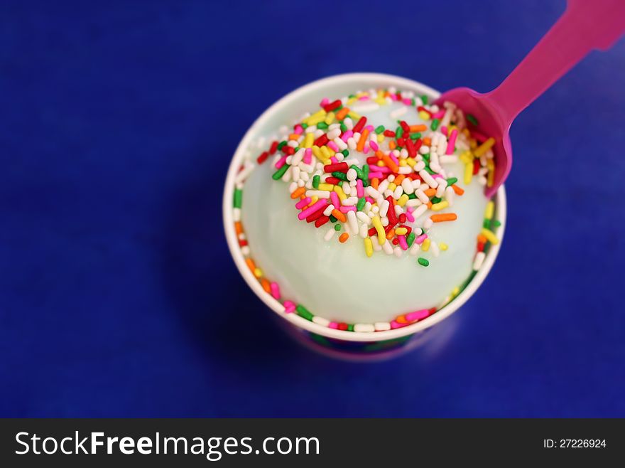 Ice-cream in a cup with a pink spoon, topped with colorful sprinkles, on a bright blue table top background. Ice-cream in a cup with a pink spoon, topped with colorful sprinkles, on a bright blue table top background