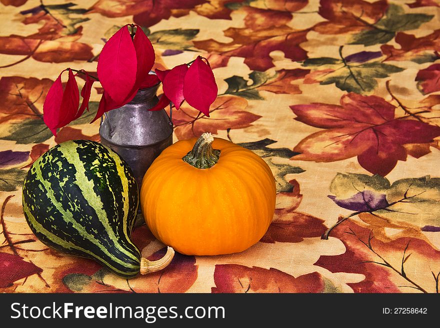 Miniature antique milk can with red leaves from a burning bush, a striped gourd, and a tiny pumpkin assembled as a centerpiece on an autumn tablecloth. Miniature antique milk can with red leaves from a burning bush, a striped gourd, and a tiny pumpkin assembled as a centerpiece on an autumn tablecloth