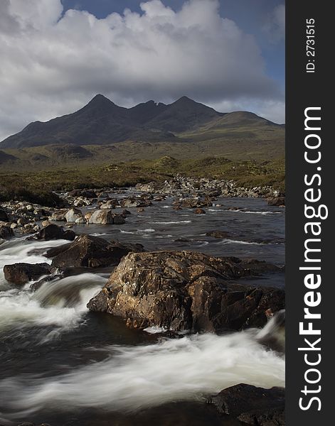 Morning sunlight lights the distant cullin hills in a warm glow. The photo is taken over a raging mountain stream with large rocks dominating the foreground. Morning sunlight lights the distant cullin hills in a warm glow. The photo is taken over a raging mountain stream with large rocks dominating the foreground