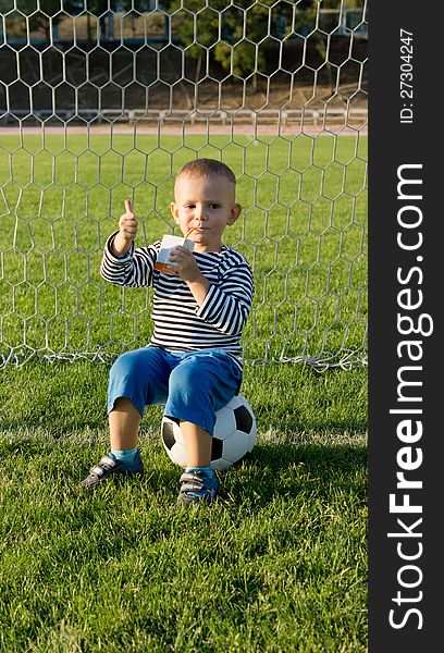 Small boy sitting on a soccer ball in a goalpost giving a thumbs up sign of approval with his hand. Small boy sitting on a soccer ball in a goalpost giving a thumbs up sign of approval with his hand