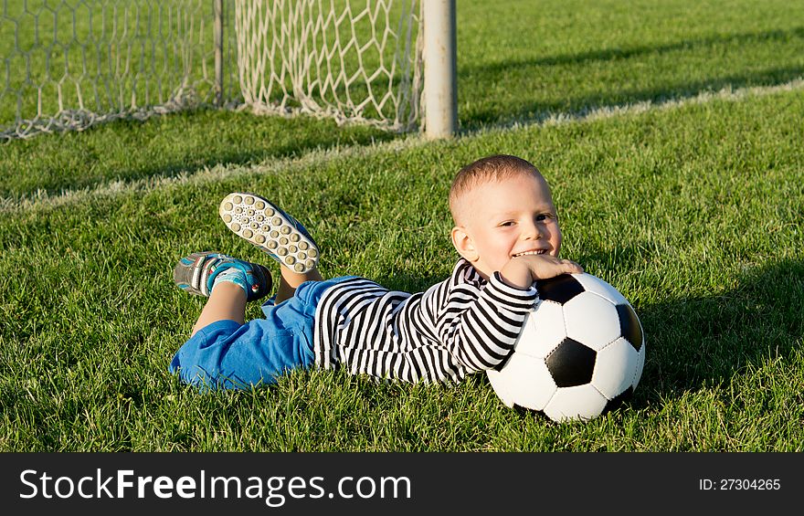 Happy little boy with his soccer ball lying on the lush green grass of a summer sportsfield in evening sunlight