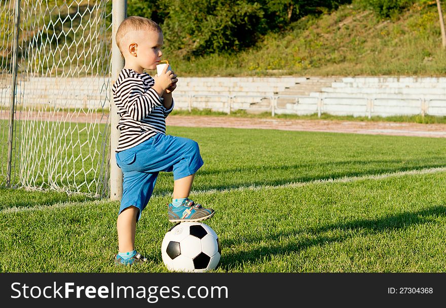 Little boy soccer player standing with hisfoot on the ball drinking juice in evening light as he takes a break from playing. Little boy soccer player standing with hisfoot on the ball drinking juice in evening light as he takes a break from playing