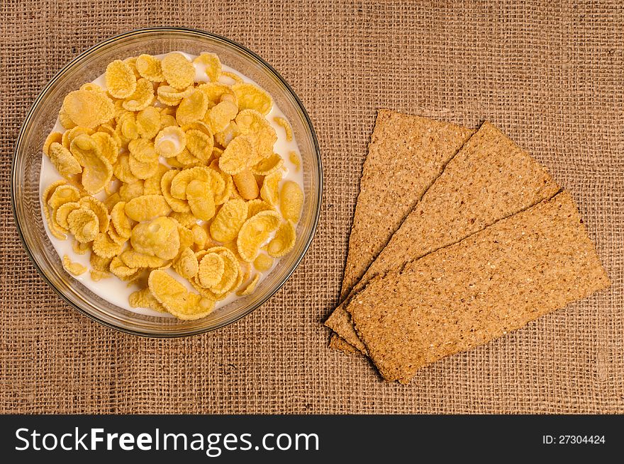 Bowl of cornflakes with milk and crispbreads on sacking background