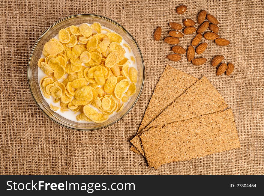 Bowl of cornflakes with milk, crispbreads and almonds on sacking background