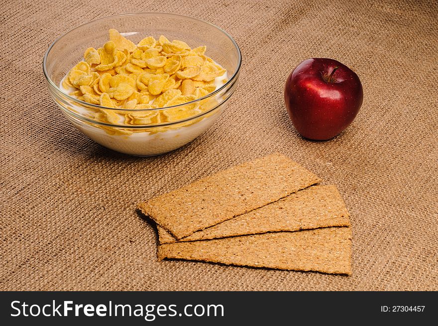 Bowl of cornflakes with milk, crispbreads and apple on sacking background