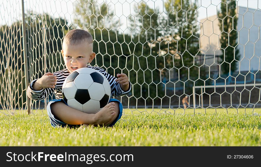 Little boy sitting cross legged on the ground in the goalposts with a soccer ball balanced on his lap. Little boy sitting cross legged on the ground in the goalposts with a soccer ball balanced on his lap