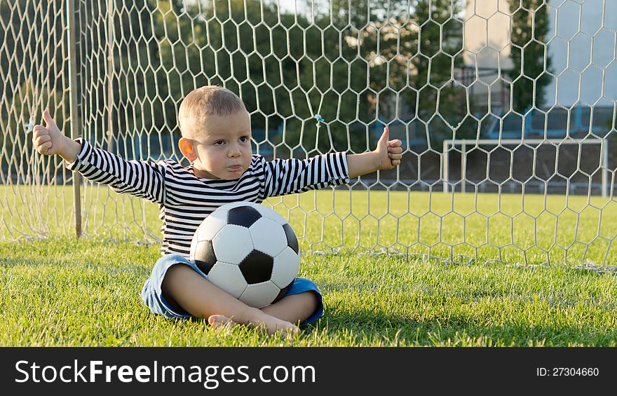 Little sportsman sitting cross lgged on green grass in the goalpost with a soccer ball on his lap giving a thumbs up. Little sportsman sitting cross lgged on green grass in the goalpost with a soccer ball on his lap giving a thumbs up