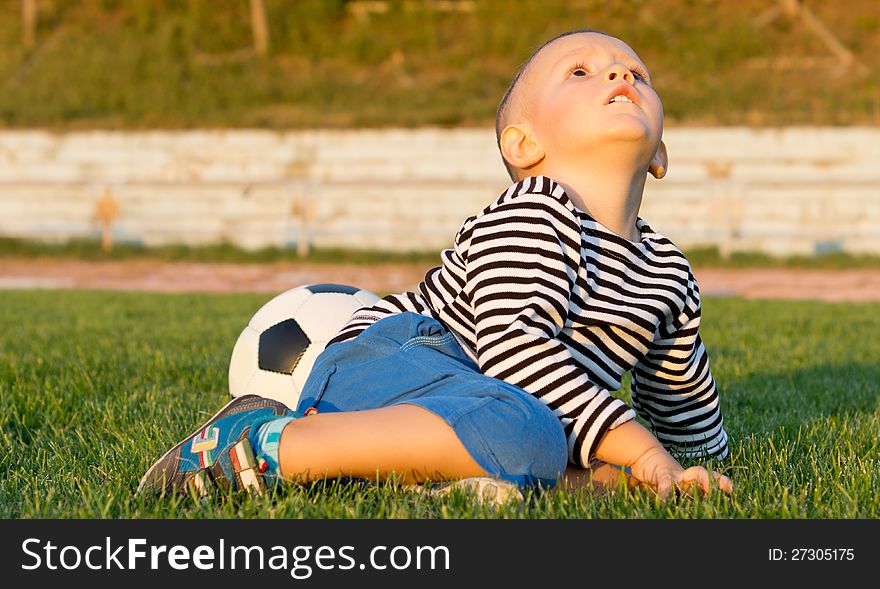 Little boy sitting on green grass with his soccer ball in evening light looking up at the sky. Little boy sitting on green grass with his soccer ball in evening light looking up at the sky