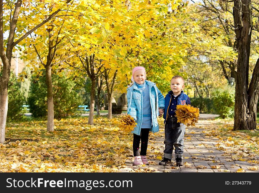 Small young brother and sister standing together on a walkway under trees in a colourful autumn park with handfuls of yellow fall leaves. Small young brother and sister standing together on a walkway under trees in a colourful autumn park with handfuls of yellow fall leaves