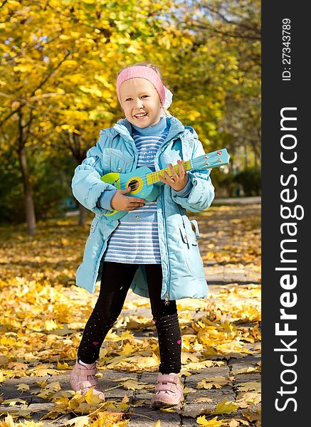 Cute little girl with a happy grin standing outdoors in an autumn park playing a toy guitar. Cute little girl with a happy grin standing outdoors in an autumn park playing a toy guitar