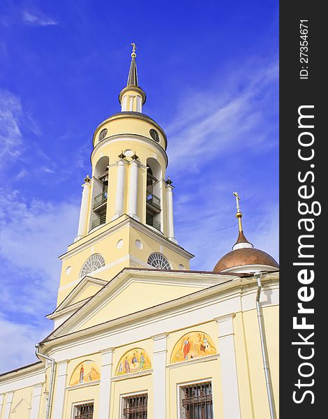 Photo of bell tower of yellow church with hours, cross, arches and columns, bells against blue morning sky. Photo of bell tower of yellow church with hours, cross, arches and columns, bells against blue morning sky