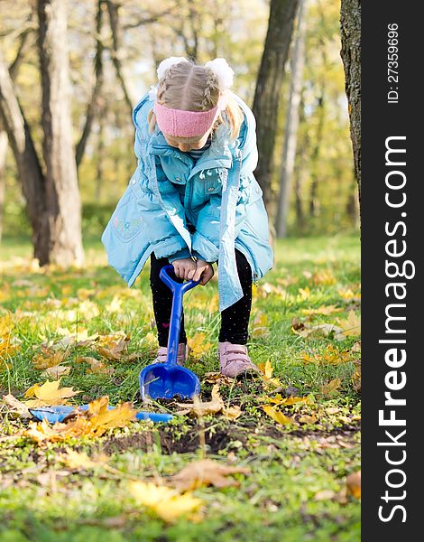 Little blonde girl in warm autumn clothing digging in a park with woodland trees. Little blonde girl in warm autumn clothing digging in a park with woodland trees