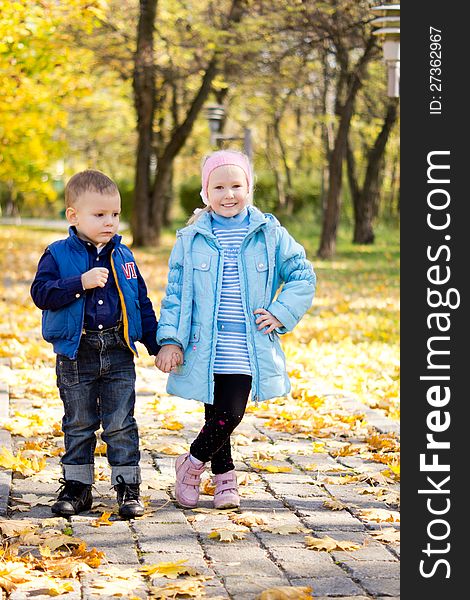 Young brother and sister holding hands and standing posing on a paved walkway in an autumn park with colouful yellow leaves. Young brother and sister holding hands and standing posing on a paved walkway in an autumn park with colouful yellow leaves