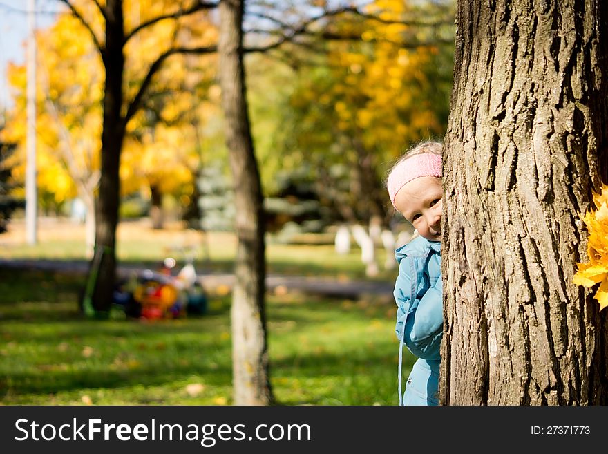 Young girl playing peek a boo hiding behimd the trunk of tree in an autumn park and peering out cheekily from the side
