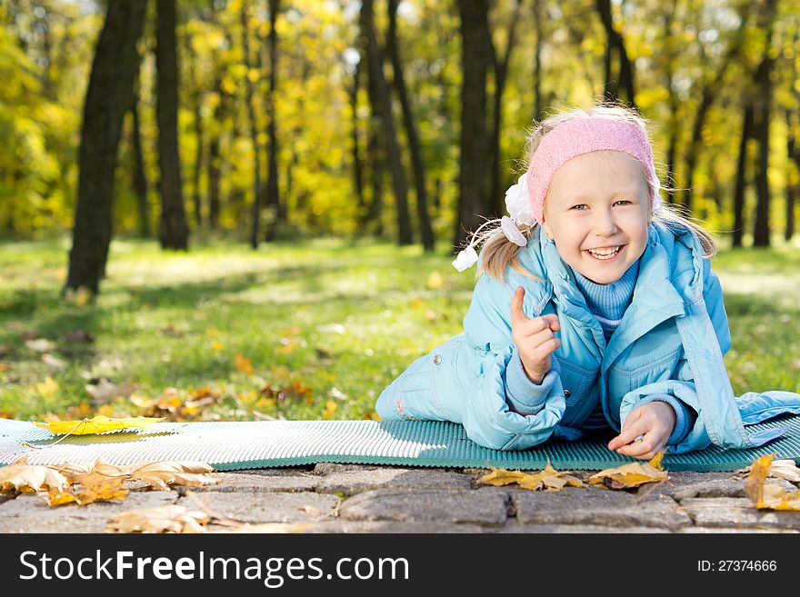 Laughing little girl lying on the ground on a mat in woodland pointing at the camera with her finger. Laughing little girl lying on the ground on a mat in woodland pointing at the camera with her finger