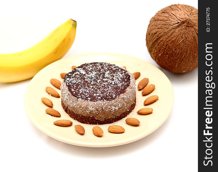 Home made cake on a brown plate covered with chocolate cream and coconut flakes on a white table with banana, almond seeds and coconut. Home made cake on a brown plate covered with chocolate cream and coconut flakes on a white table with banana, almond seeds and coconut