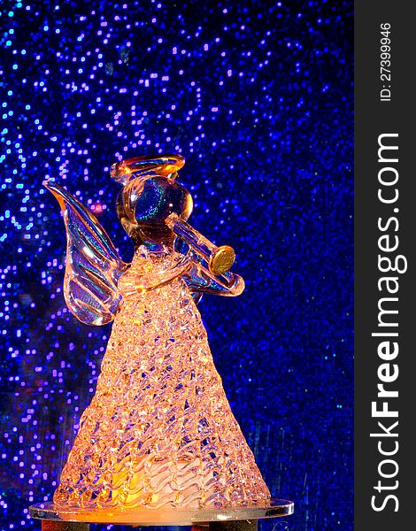 A statuette consistin of glass representing an angel which blows a trumpet. Background blue with glittering spots like stars, the glass is lit from behind with orange colored light. A statuette consistin of glass representing an angel which blows a trumpet. Background blue with glittering spots like stars, the glass is lit from behind with orange colored light