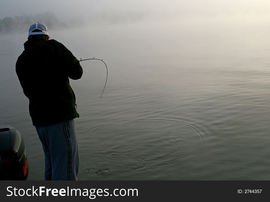 Angler fishing in a boat with a fish on and fog in the background. Angler fishing in a boat with a fish on and fog in the background.
