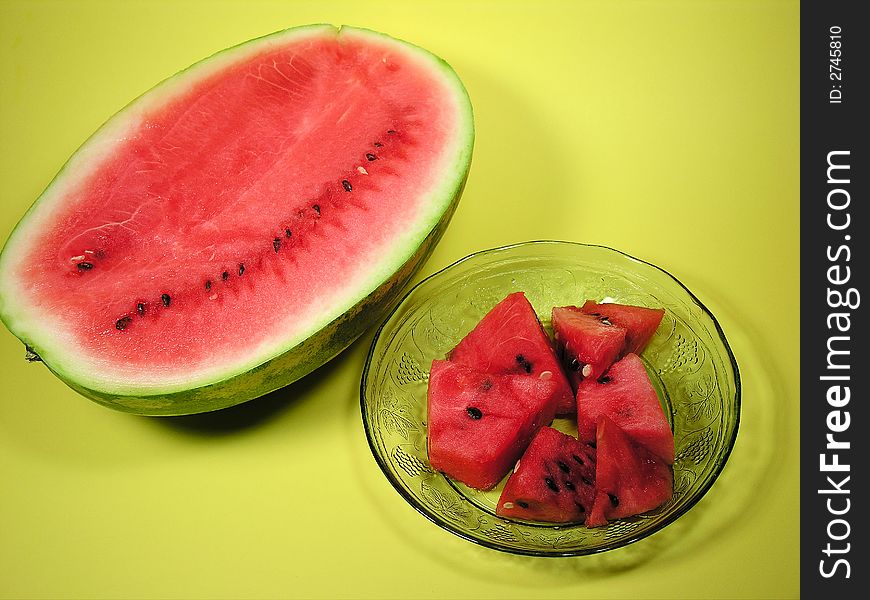 One half of a large cut watermelon beside a pretty crystal dish with fruit designs engraved into the glass, filled with fresh red watermelon pieces against a yellow background. One half of a large cut watermelon beside a pretty crystal dish with fruit designs engraved into the glass, filled with fresh red watermelon pieces against a yellow background.